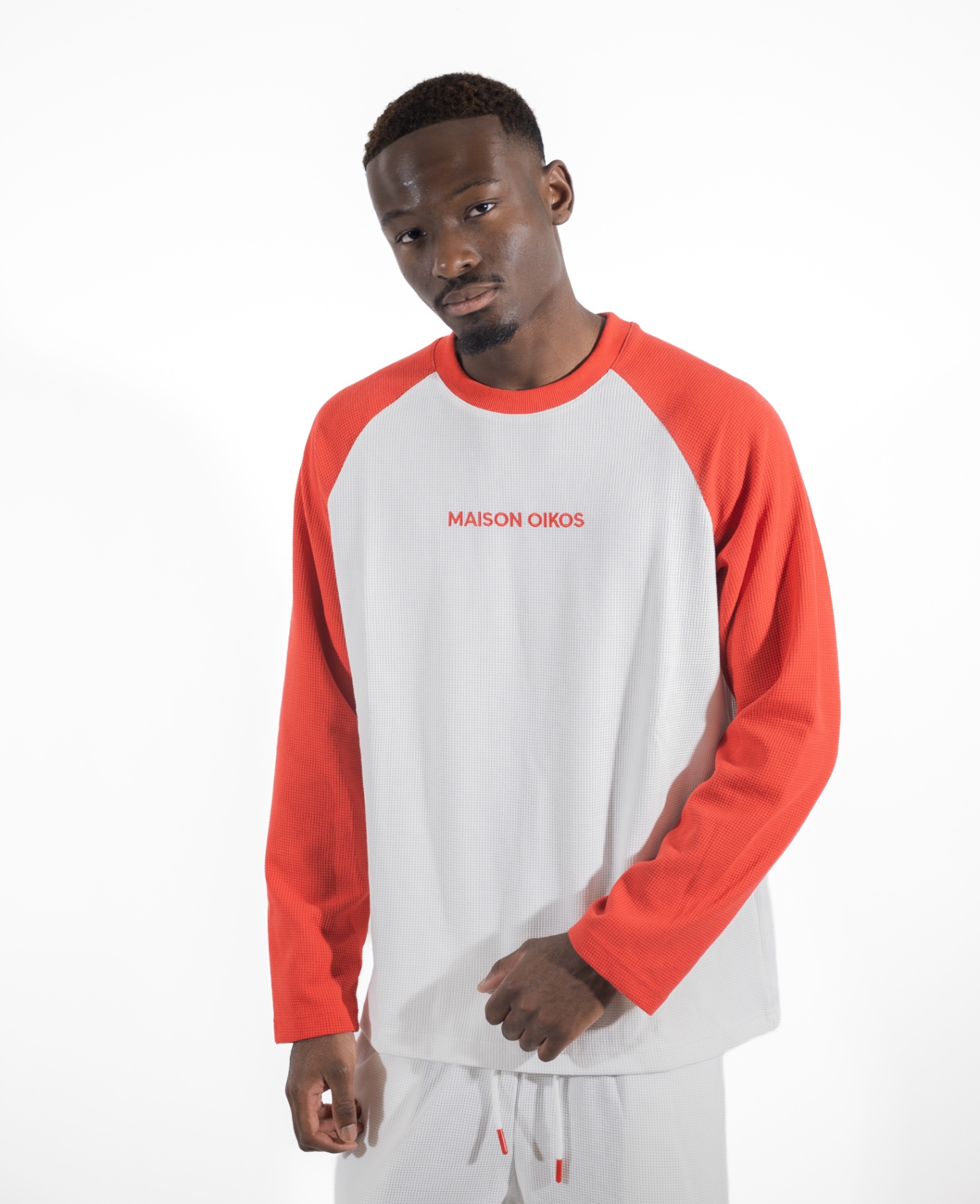 Homme de face en haut raglan blanc à manches rouges et short blanc, logo Maison Oikos en rouge.