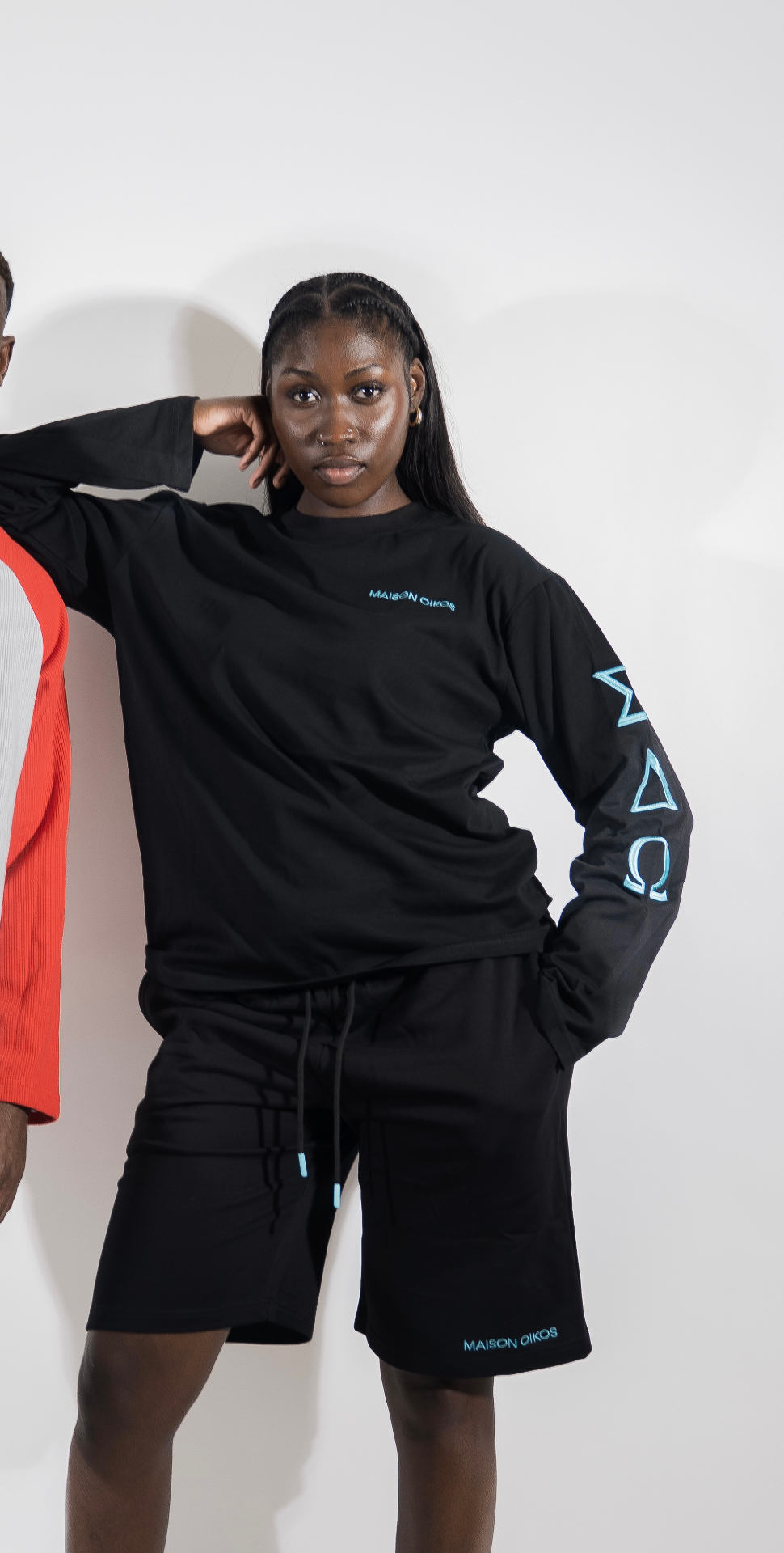 Femme de face en ensemble noir Maison Oikos : haut à manches longues et short assorti, logo bleu ciel sur la poitrine.