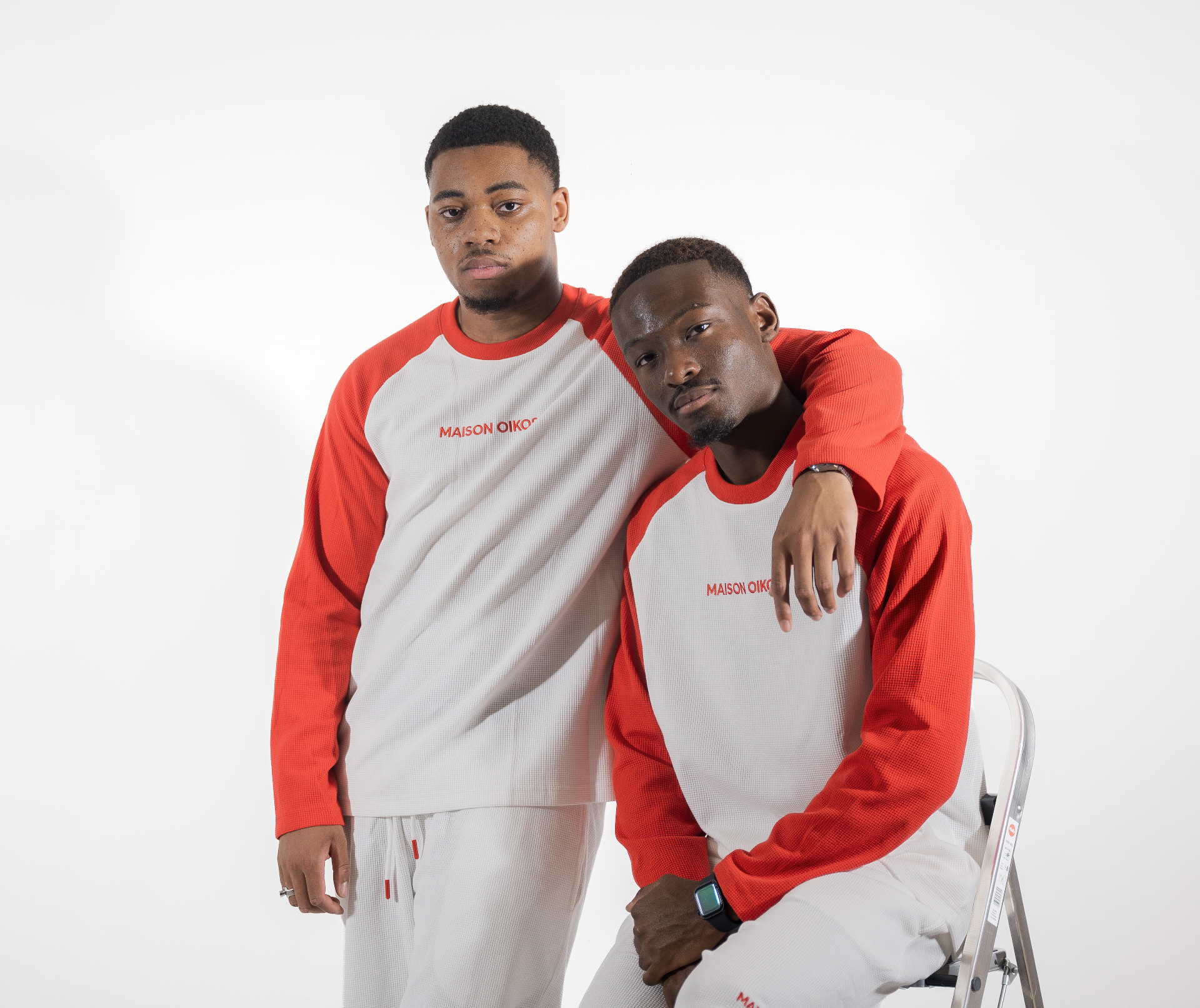 Deux hommes en studio, l'un debout l'autre assis, en haut raglan blanc et rouge et short blanc Maison Oikos assorti.