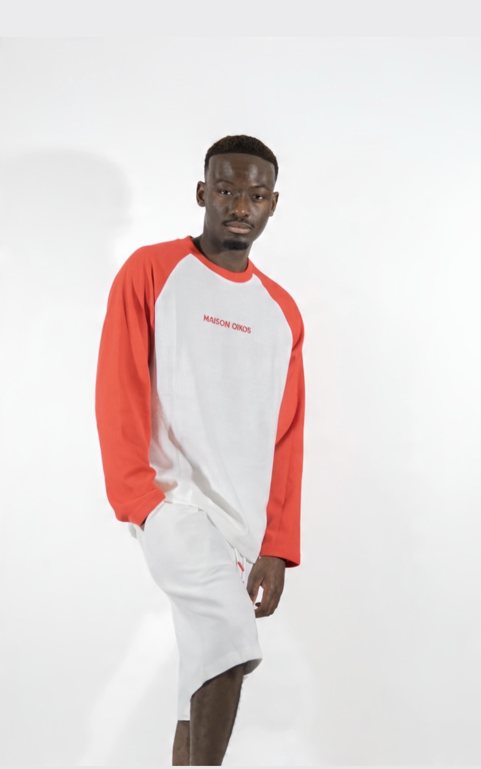 Homme debout en ensemble complet : haut raglan blanc et rouge avec short blanc Maison Oikos assorti.